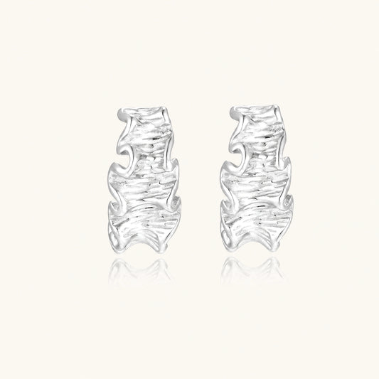 Textured white gold-plated hoop earrings with flowing wave pattern against cream background, showing dimensional rippled surface.