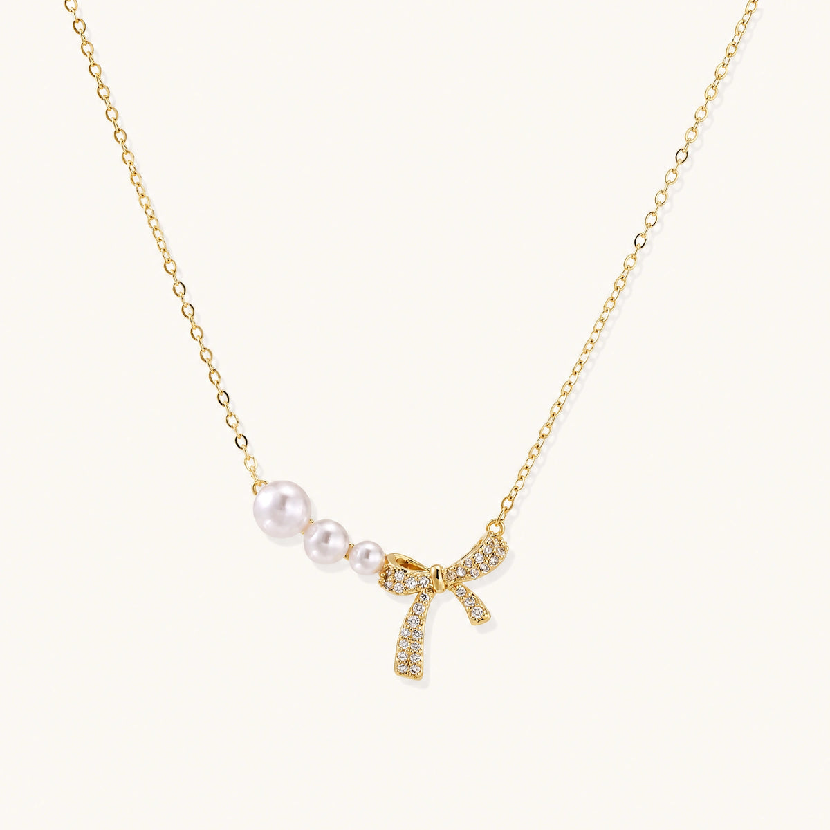 Delicate gold necklace with three white pearls and crystal-embellished bow pendant on cream background