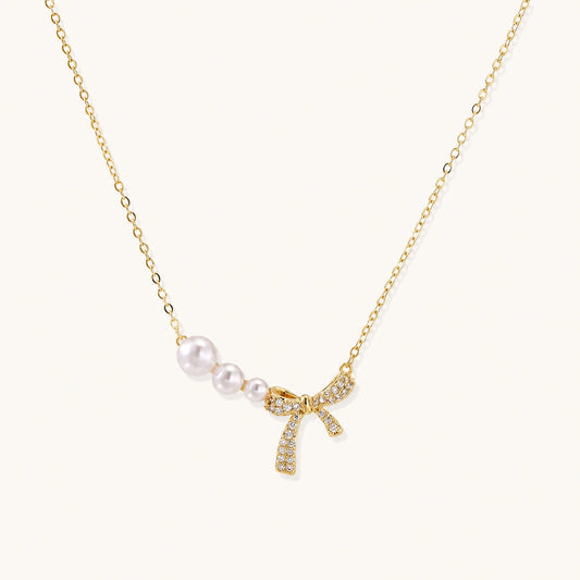 Delicate gold necklace with three white pearls and crystal-embellished bow pendant on cream background