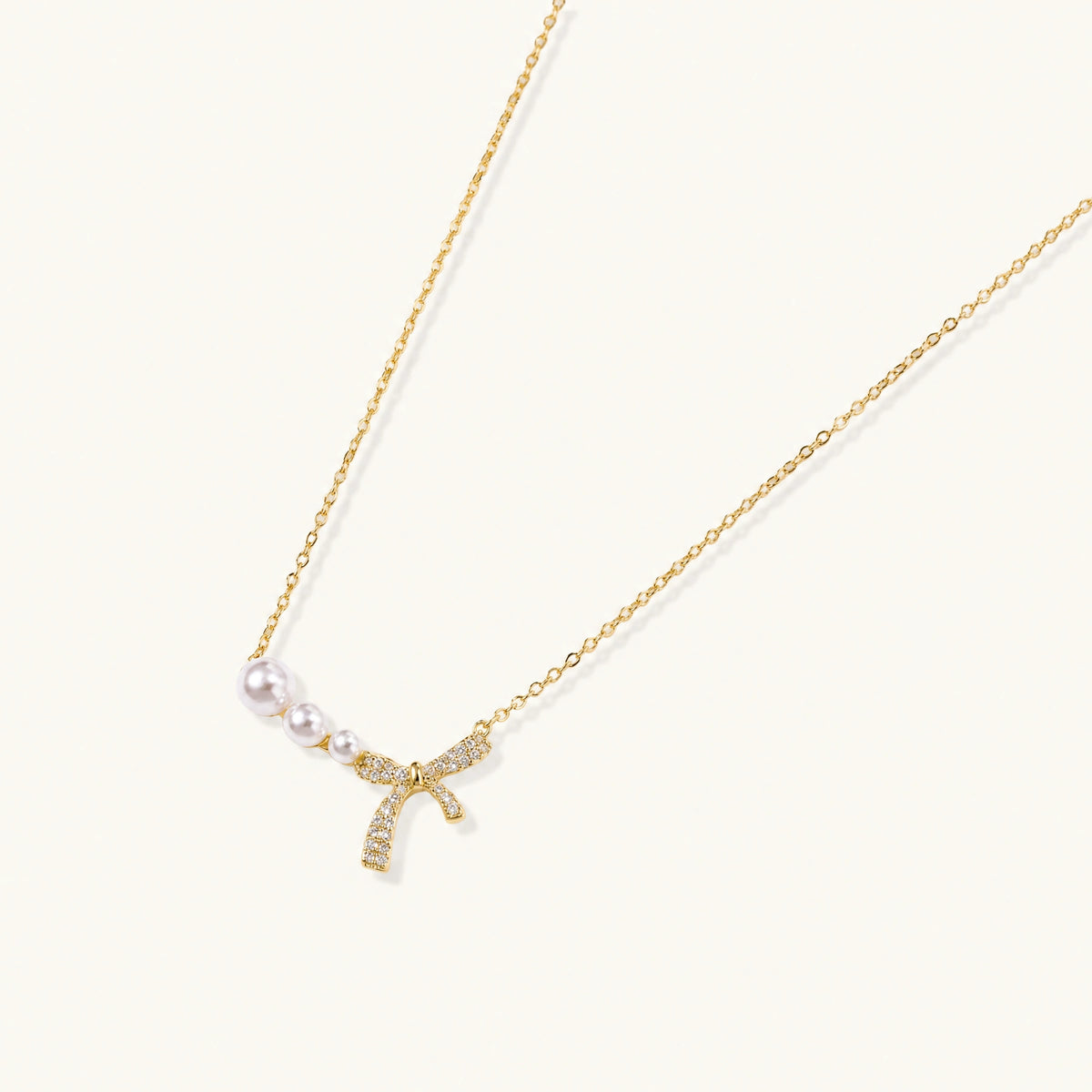 Delicate gold necklace with three white pearls and crystal-embellished bow pendant on cream background