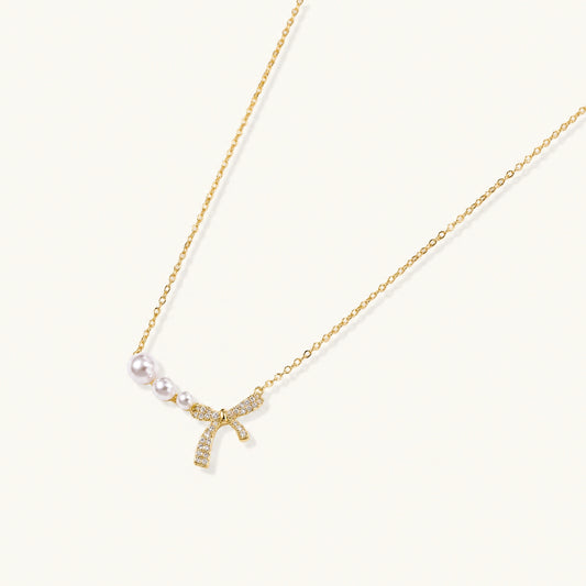 Delicate gold necklace with three white pearls and crystal-embellished bow pendant on cream background