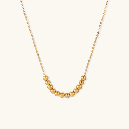 Delicate gold-plated necklace with graduated spherical beads forming gentle smile curve on beige background