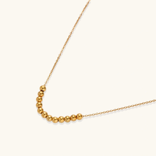 Delicate gold-plated necklace with graduated spherical beads forming gentle smile curve on beige background
