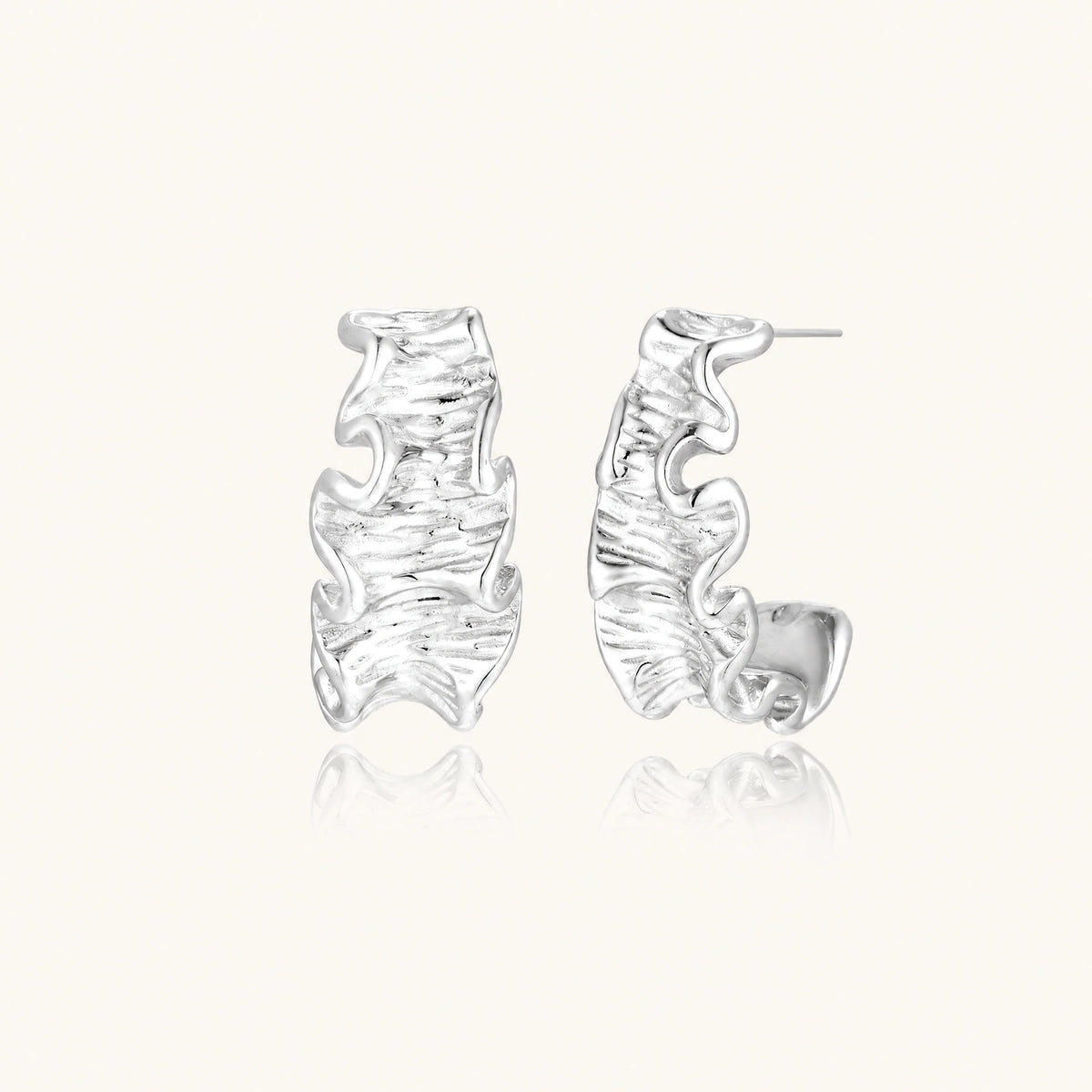 Textured white gold-plated hoop earrings with flowing wave pattern against cream background, showing dimensional rippled surface.