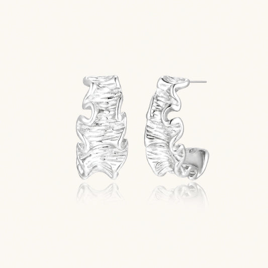 Textured white gold-plated hoop earrings with flowing wave pattern against cream background, showing dimensional rippled surface.