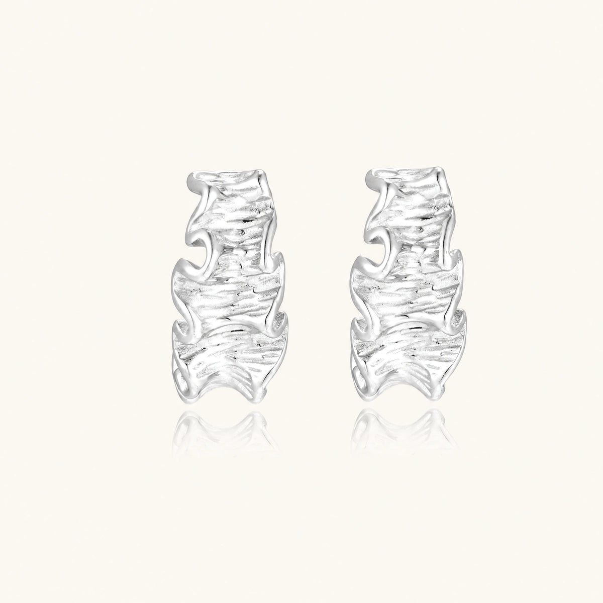 Textured white gold-plated hoop earrings with flowing wave pattern against cream background, showing dimensional rippled surface.