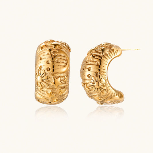 Wide gold hoop earrings with embossed stars and sun motifs on cream background, showing dimensional relief details.