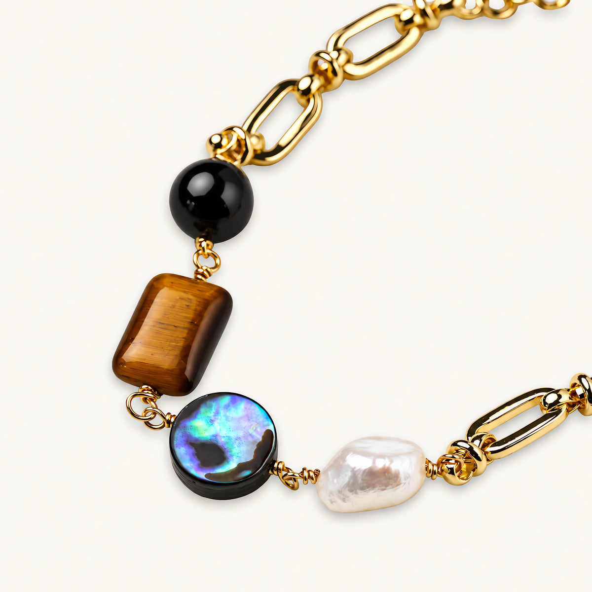 Gold chain bracelet with tiger's eye, obsidian, pearl, and iridescent abalone stones on cream background.